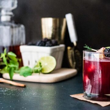 A dark red cocktail bursting with berry flavor is garnished with blackberries and a sprig of mint, sitting elegantly on a table. Behind it, a wooden board holds a lime and blackberries, alongside a decanter and gold shaker, while a small wooden spoon rests nearby.