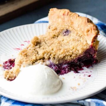 A slice of berry crumble pie is served on a white plate with a scoop of vanilla ice cream, epitomizing fall comfort food. The plate sits on a blue and white striped napkin, and the filling appears to be leaking onto the plate. The background includes a wooden cutting board.