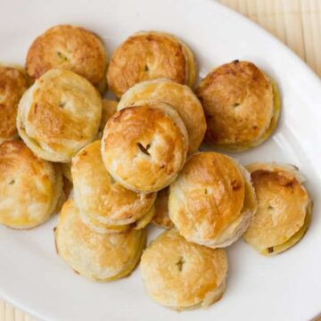 A white oval plate holds several round, golden-brown pastries. The pastries are stacked unevenly, with slight variations in size. Each pastry has a small indentation in the center. This trending recipe features popular treats on a light-colored, textured surface.