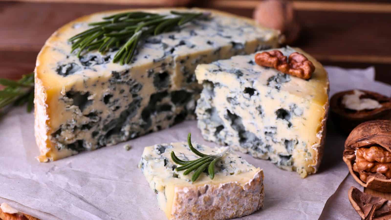 A wheel of blue cheese sits on parchment paper, with a wedge cut out, displaying its blue veins. The cheese is garnished with sprigs of rosemary, and there are scattered walnuts around it. The background is a wooden surface.