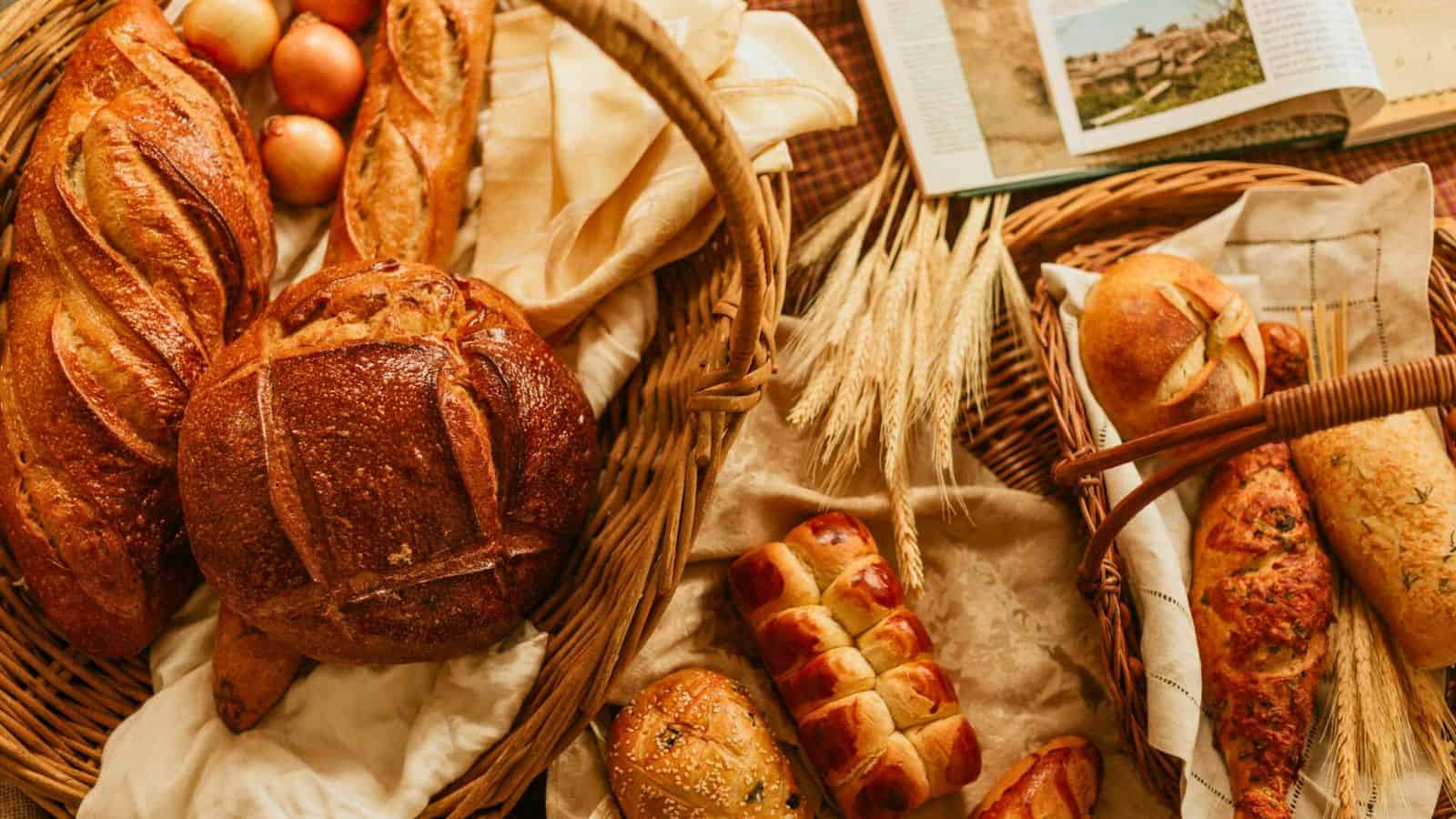 A variety of bread and rolls arranged in baskets, accompanied by wheat stalks, apples, and an open book. The breads have different shapes and textures, including round loaves, braided rolls, and baguettes, set on a rustic tablecloth.