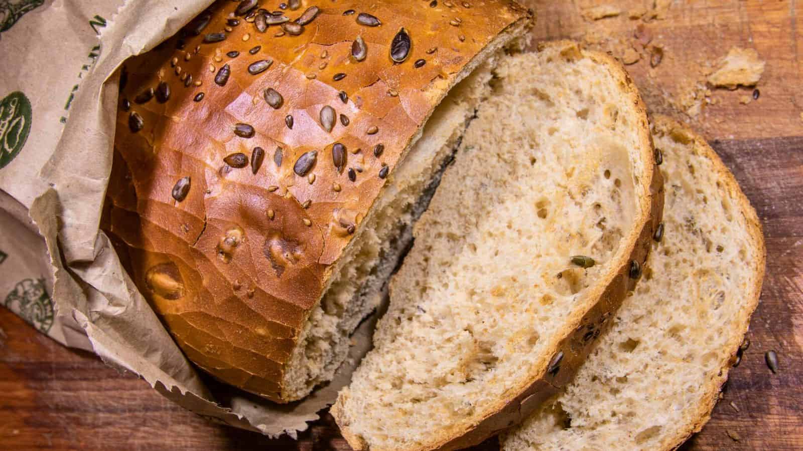 A loaf of seeded bread partially wrapped in paper is placed on a wooden surface. Two slices of the bread have been cut and are positioned in front of the loaf, showing the textured interior with seeds scattered throughout.