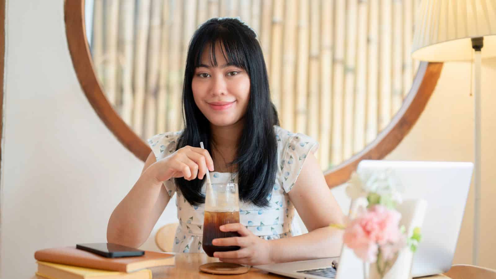 A person with long black hair is seated at a wooden table, holding a glass of iced coffee and stirring it with a straw. A laptop, a phone, and some books are on the table. There is a round window and a lamp in the background.