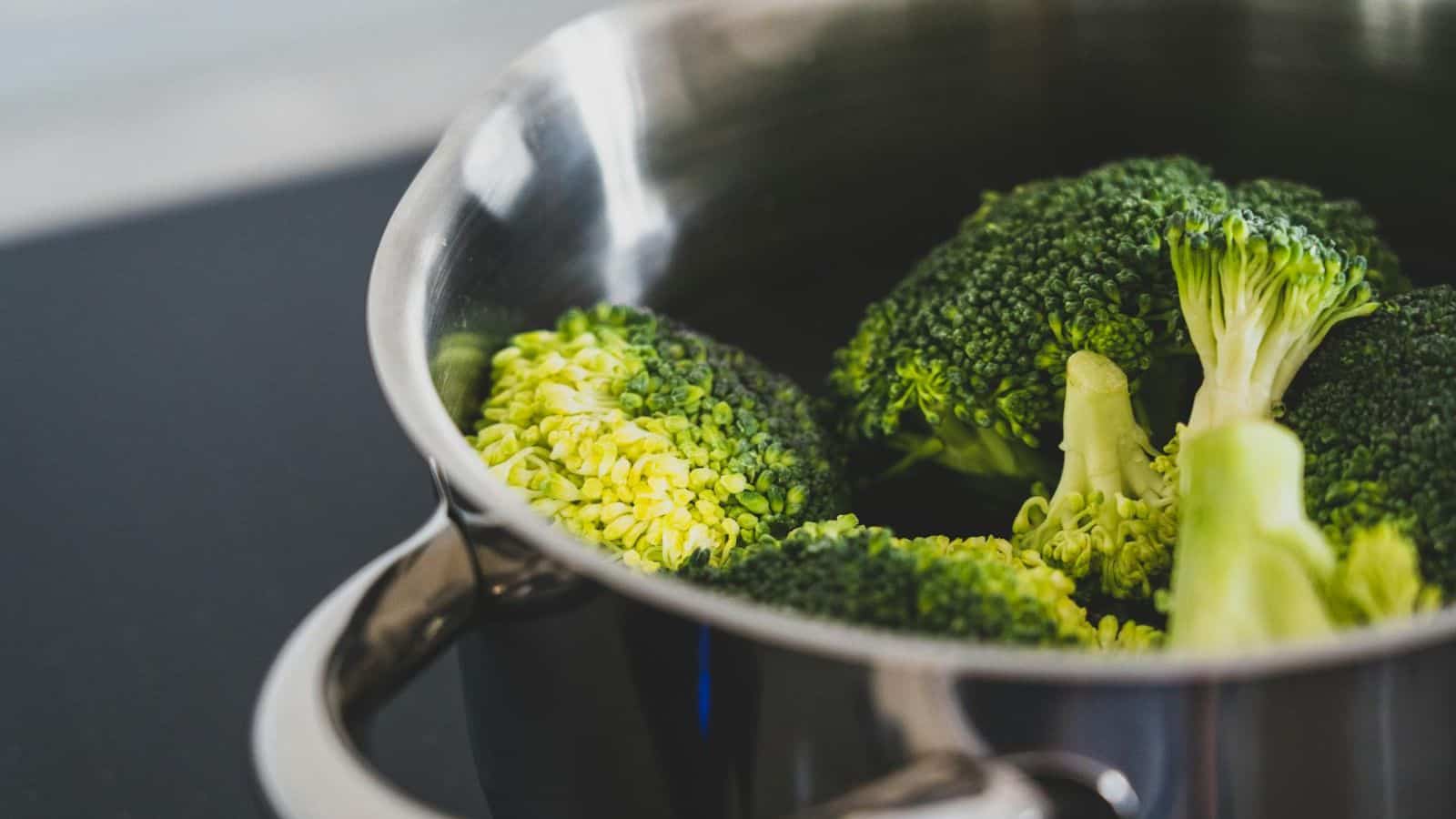 A close-up of broccoli florets being steamed in a stainless steel pot. The bright green vegetables are partially submerged in water, and the pot is placed on a flat, black surface.