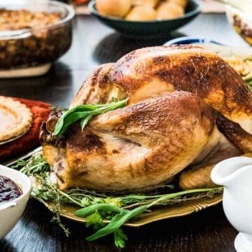 A roasted turkey garnished with herbs is placed at the center of a dining table surrounded by various dishes, including cranberry sauce, pie, green beans, stuffing, and a gravy boat—a feast filled with Thanksgiving-worthy recipes to wow everyone.