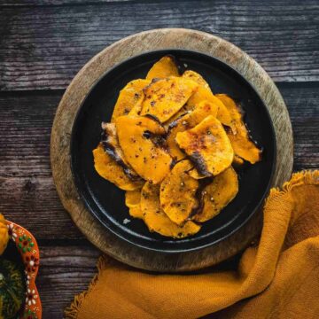 A black bowl filled with baked butternut squash slices seasoned with spices is placed on a wooden cutting board on a dark wooden surface. To the left, there is a ceramic bowl with ornamental gourds, and to the right, there is an orange napkin. This dish highlights trending recipes perfect for any cozy dinner setup.