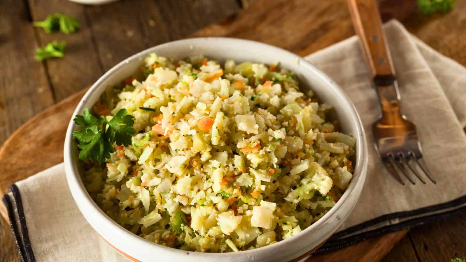 A white bowl filled with cauliflower rice mixed with diced vegetables, placed on a cloth napkin with a fork beside it. The dish is garnished with a parsley leaf. The background includes a wooden surface and some scattered greens.