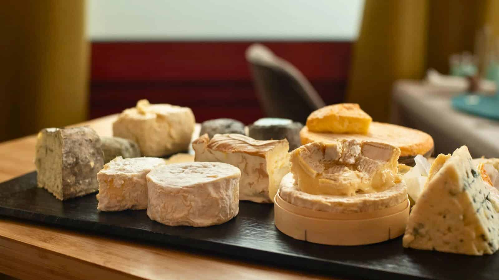 A display of various types of cheese is arranged on a dark serving board set on a wooden table. The cheeses vary in shape, size, and texture, including round, wedge-shaped, and wheel forms, some with rinds and others with creamy interiors.