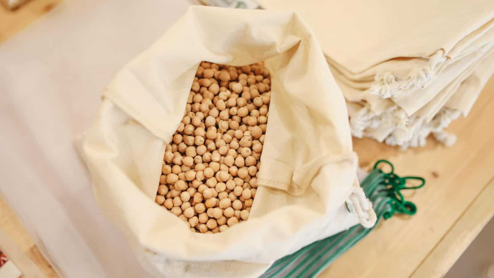 A cloth bag filled with chickpeas sits on a table. Adjacent to the bag are stacked white cloth bags and a few green plastic clips. The scene appears to be at a store, likely in a bulk food section.