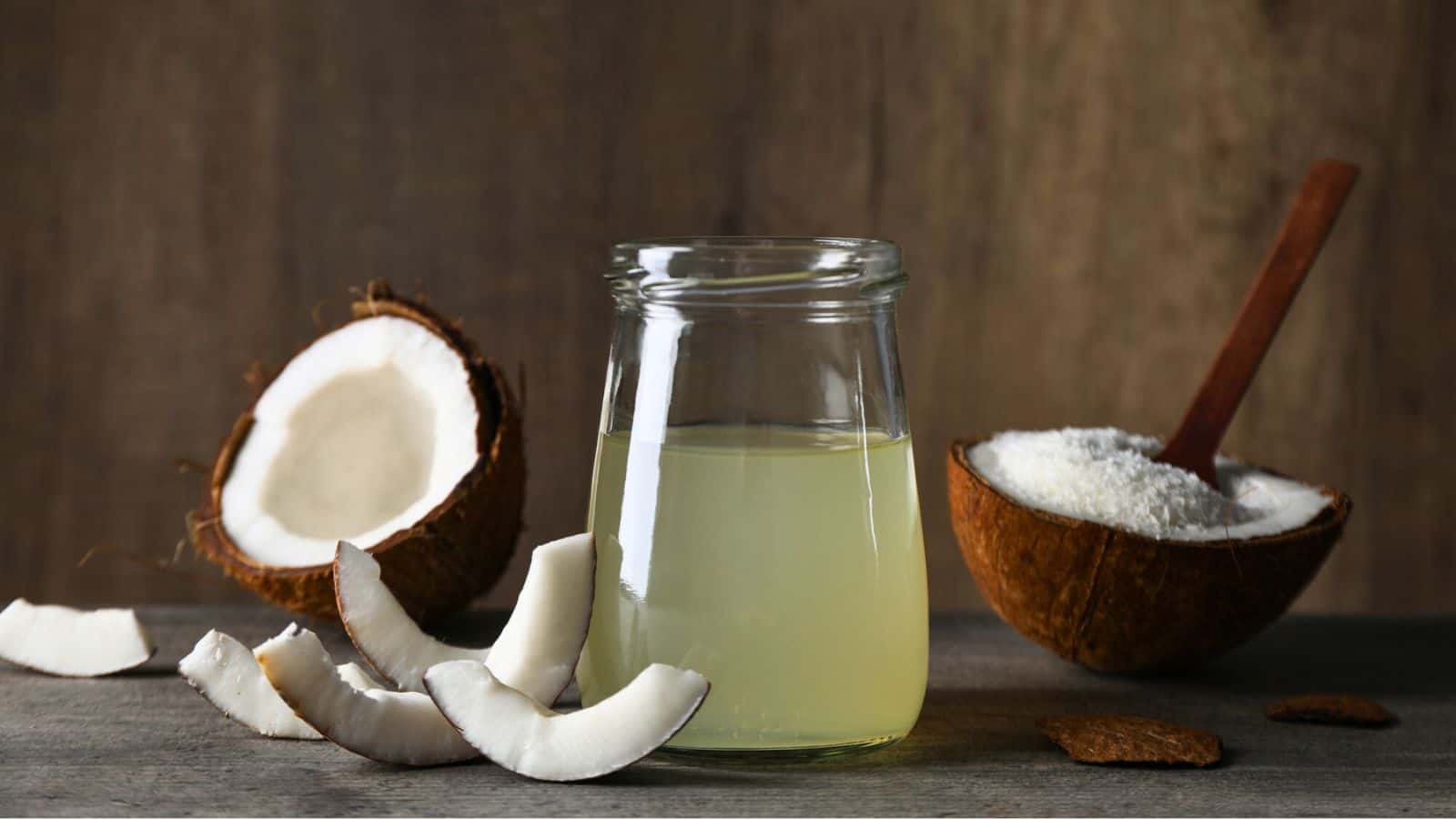 A glass jar filled with coconut water is placed on a wooden surface. Next to it, there are coconut halves and several coconut pieces. A wooden spoon rests in one of the coconut halves containing coconut flour. The background is a blurred wooden texture.
