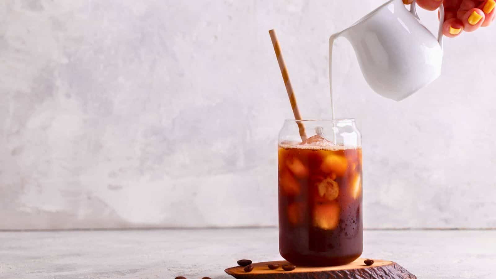 A glass of iced coffee with a straw sits on a wooden coaster next to several coffee beans. A hand is pouring milk from a small white pitcher into the glass. The background is a light, neutral color.