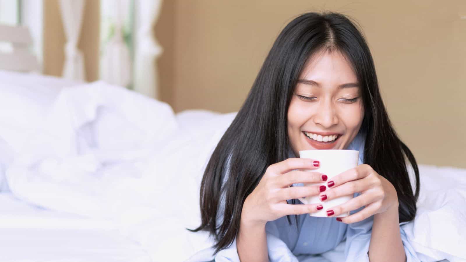 A person with long black hair is lying on a bed, holding a white mug close to their face and smiling. The bed has white sheets and is in a room with light-colored walls and sheer curtains by the window.