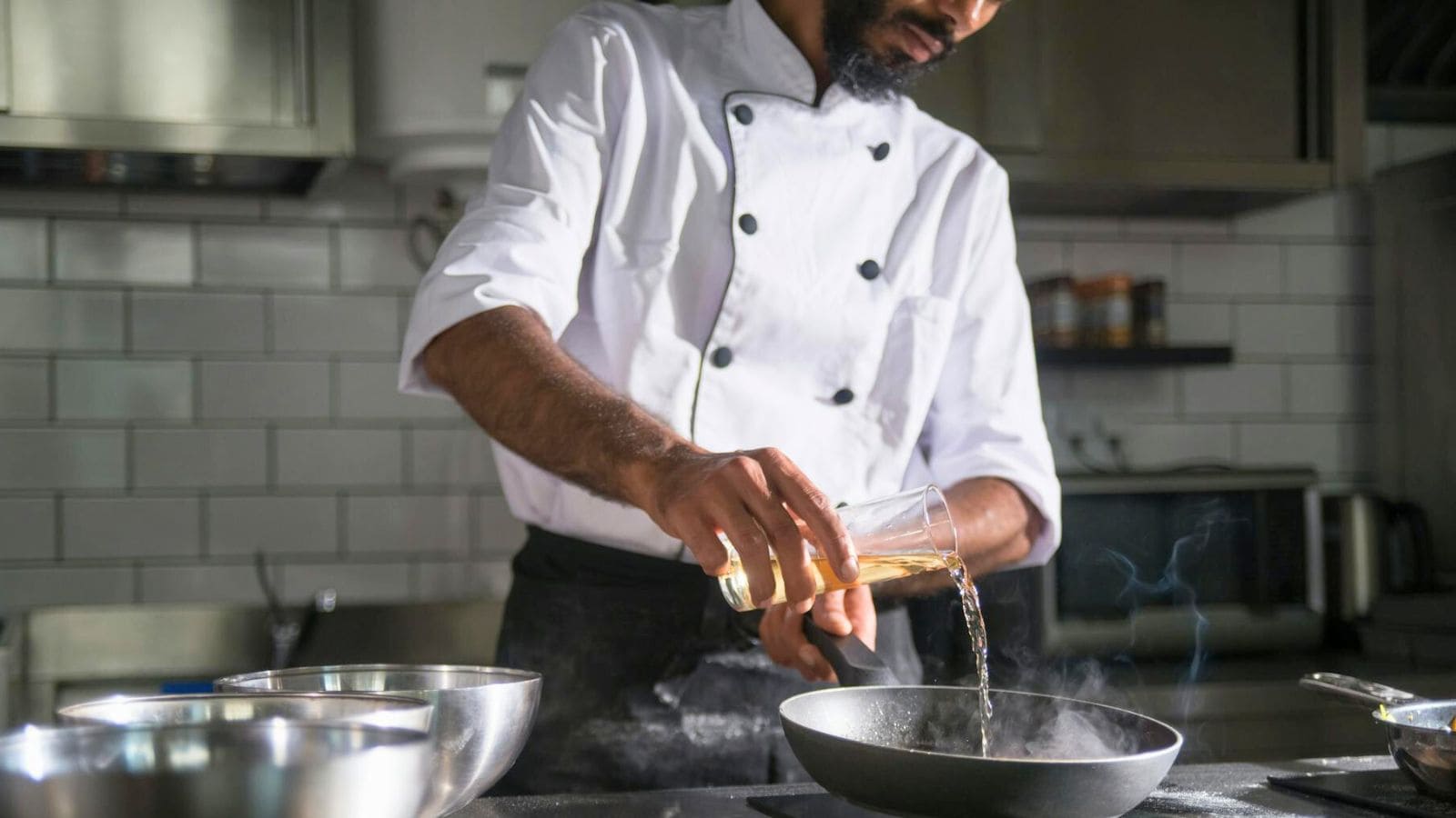 A chef, dressed in a white double-breasted jacket, pours liquid from a glass into a frying pan. The kitchen is equipped with a white-tiled backsplash, metallic mixing bowls, and various kitchen utensils.