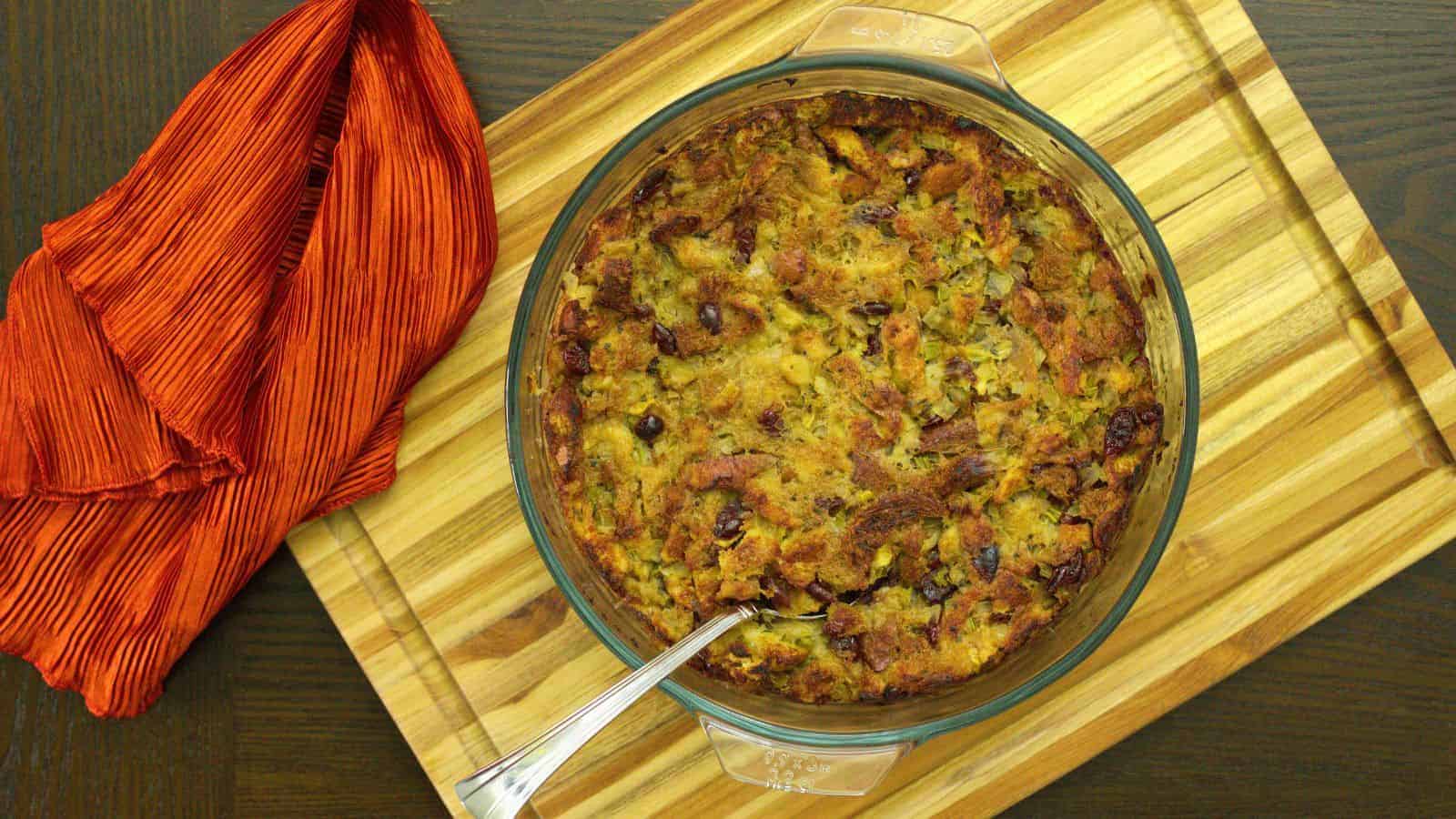 A round, clear glass baking dish filled with baked stuffing sits on a wooden cutting board. A silver serving spoon rests inside the dish. To the left, there is a folded red-striped cloth napkin. The background is a dark wooden surface, perfectly capturing the essence of fall and sweetened by seasonal cranberry recipes.