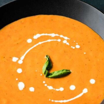 A bowl of creamy tomato soup garnished with a swirl of white cream and a few basil leaves. The black bowl, evocative of cozy fall soups, is placed on a dark surface, perfect for enjoying by the fireplace.