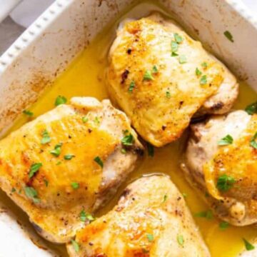 A white baking dish holds four baked chicken thighs with golden brown skin, sprinkled with chopped green herbs and sitting in a pool of juices. This healthy, low-carb dinner is placed on a white surface alongside a white cloth.