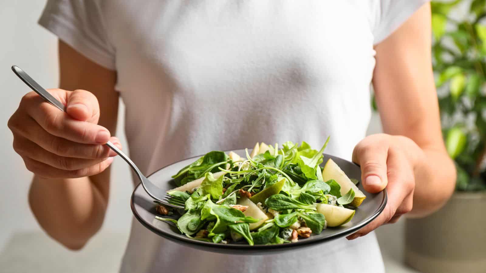 A person in a white shirt is holding a black plate with a fresh green salad that includes leafy greens, chopped vegetables, and lemon wedges. The person is holding a fork in their right hand and is about to take a bite. A plant is visible in the background.