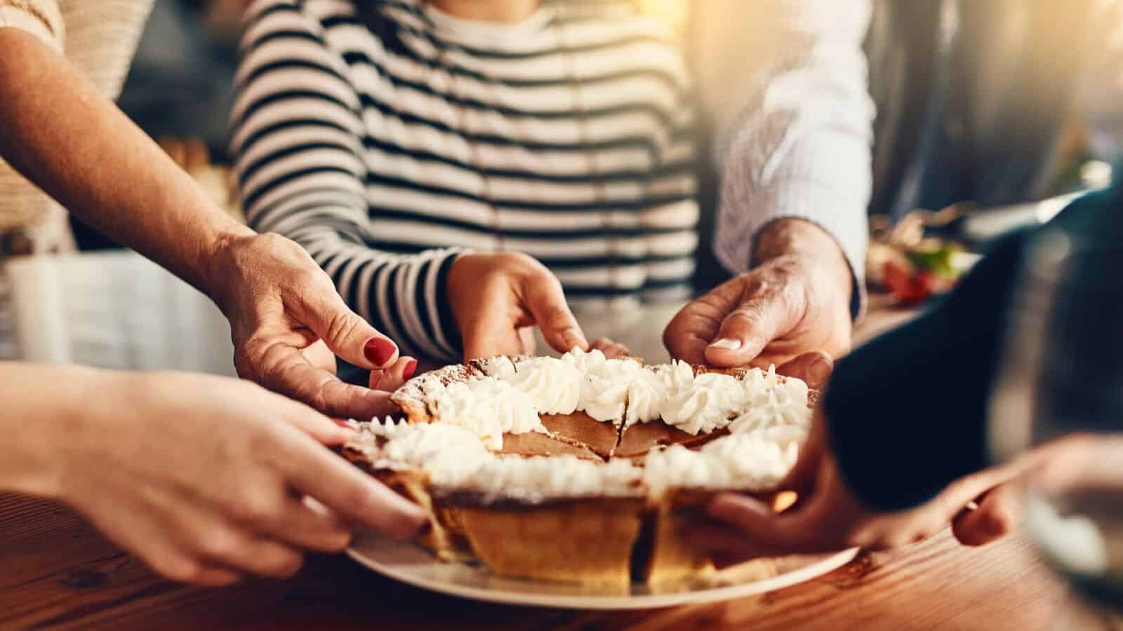 Four people are reaching out to hold or pass a plate with a whole pie decorated with whipped cream. The background includes a person wearing a striped shirt with a blurred face, and the setting appears to be casual, likely around a dining table.