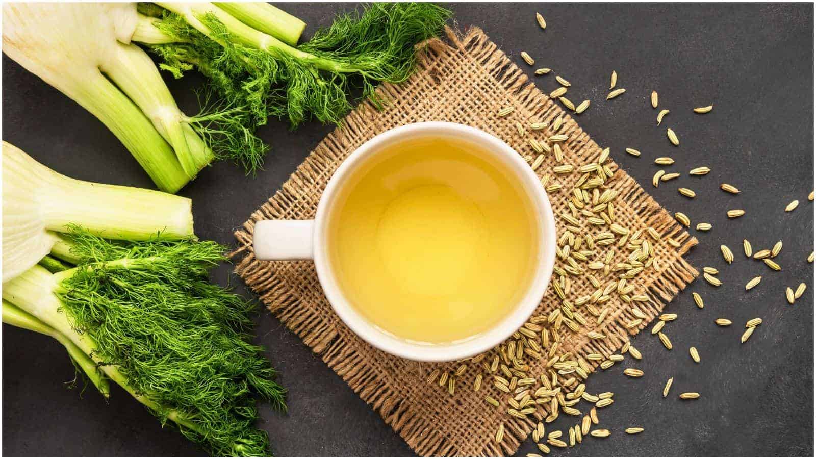 A cup of fennel tea rests on a woven mat. Fresh fennel stalks with green fronds and some scattered fennel seeds are placed around the mat on a dark surface. The tea in the cup is golden in color.