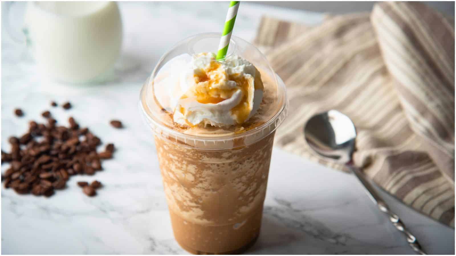 A blended iced coffee drink sits on a marble surface, topped with whipped cream and caramel drizzle, and features a green and white striped straw. A metal spoon is to the right, and coffee beans and a glass of milk are in the background.