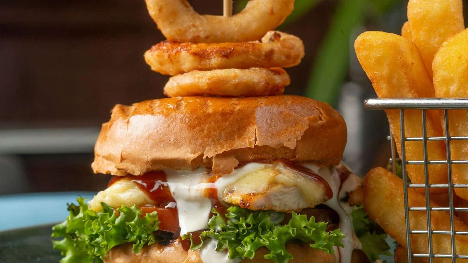 A close-up of a gourmet chicken sandwich with lettuce, melted cheese, barbecue sauce, and ranch dressing, topped with two onion rings on a wooden pick. On the right side, crispy fries are visible in a small wire basket.