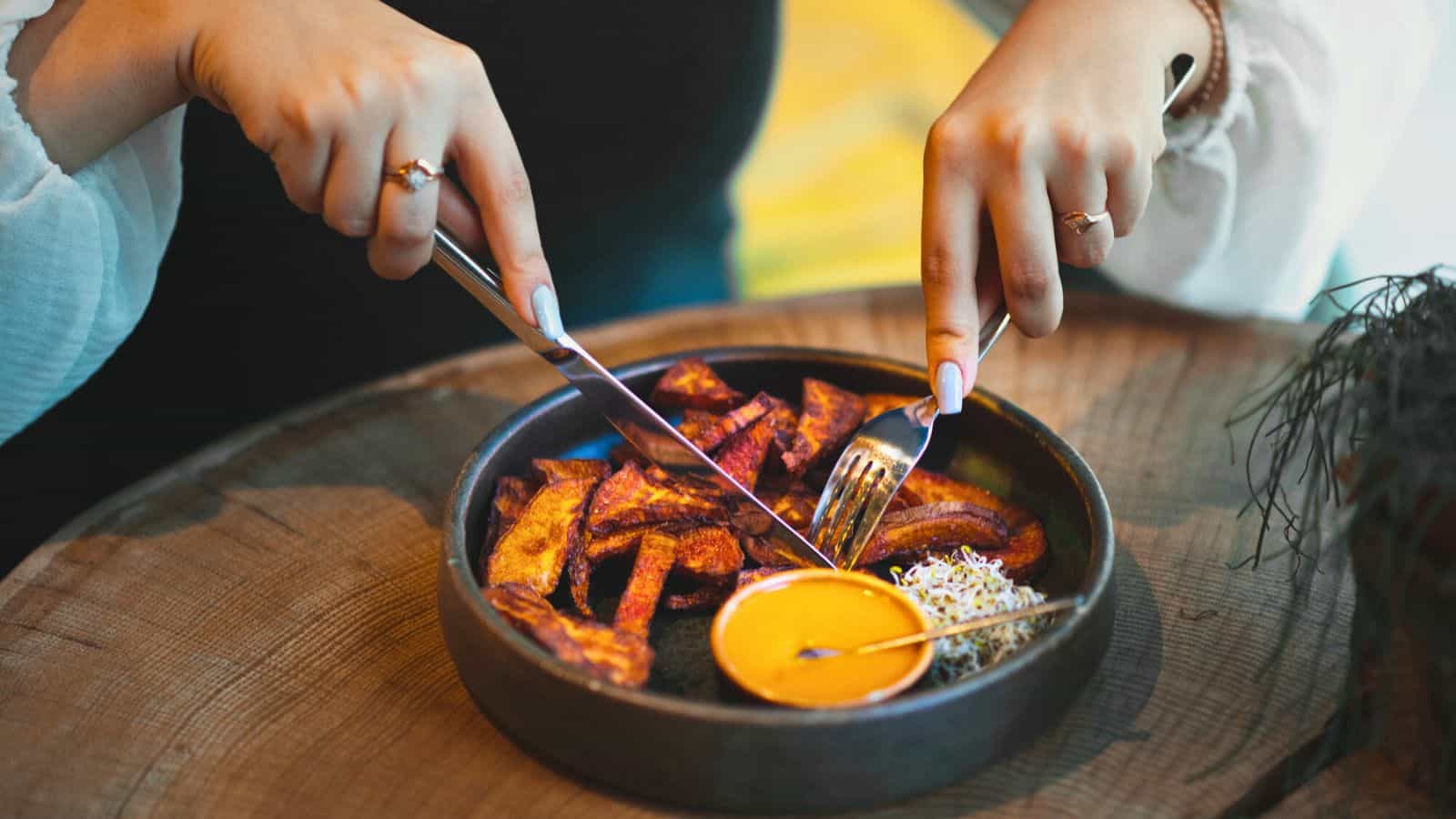 A person using a knife and fork to cut into pieces of roasted vegetables on a black plate. The plate contains an orange dipping sauce and a small serving of sprouts. The person is wearing a white long-sleeved garment. The table surface is brown wood.
