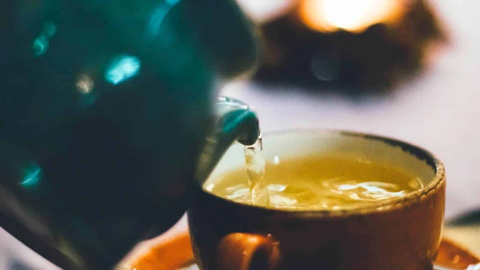 A close-up image of a liquid being poured from a green teapot into a brown ceramic cup. The liquid appears to be light-colored tea. The background is blurred with warm, soft lighting.