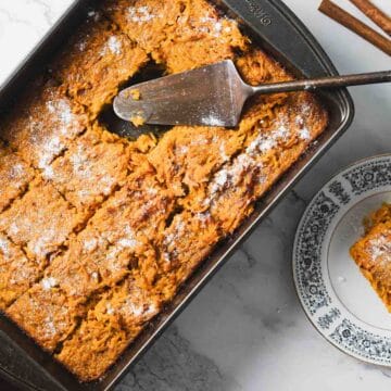 A rectangular baking dish filled with sliced baked pumpkin treat rests on a marble countertop. A slice is served on a decorative plate beside the dish. A serving spatula lies on the dish. Cinnamon sticks and whole nutmeg are scattered around.