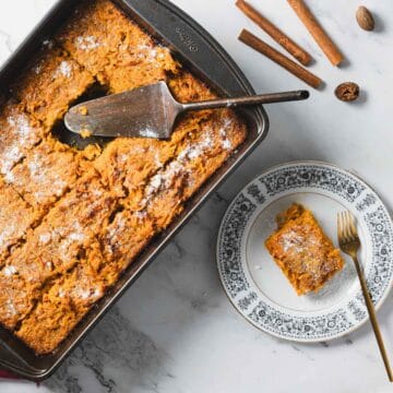 A rectangular baking dish filled with sliced baked pumpkin treat rests on a marble countertop. A slice is served on a decorative plate beside the dish. A serving spatula lies on the dish. Cinnamon sticks and whole nutmeg are scattered around.