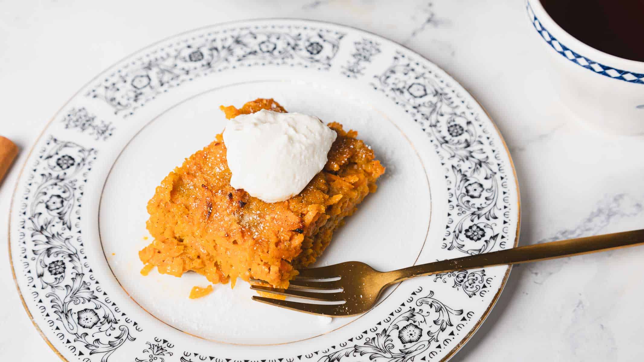 A carrot kugel slice topped with a dollop of cream sits on an ornate patterned white plate with a fork beside it. The dessert has a yellowish-orange color and is placed on a white marble surface. A black baking tray with more dessert is partially visible in the background.