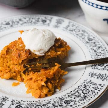 A carrot kugel slice topped with a dollop of cream sits on an ornate patterned white plate with a fork beside it. The dessert has a yellowish-orange color and is placed on a white marble surface. A black baking tray with more dessert is partially visible in the background.
