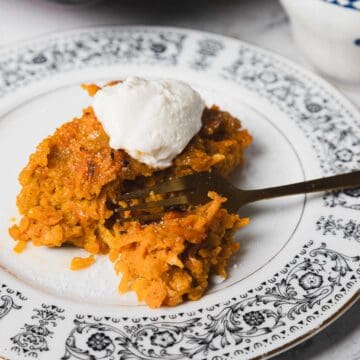 A carrot kugel slice topped with a dollop of cream sits on an ornate patterned white plate with a fork beside it. The dessert has a yellowish-orange color and is placed on a white marble surface. A black baking tray with more dessert is partially visible in the background.
