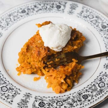 A carrot kugel slice topped with a dollop of cream sits on an ornate patterned white plate with a fork beside it. The dessert has a yellowish-orange color and is placed on a white marble surface. A black baking tray with more dessert is partially visible in the background.