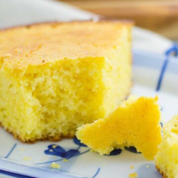 A slice of cornbread is served on a white plate with blue decorations. Two small pieces of the Auto Draft cornbread are broken off and placed next to the slice. The cornbread has a crumbly texture and a golden brown top.