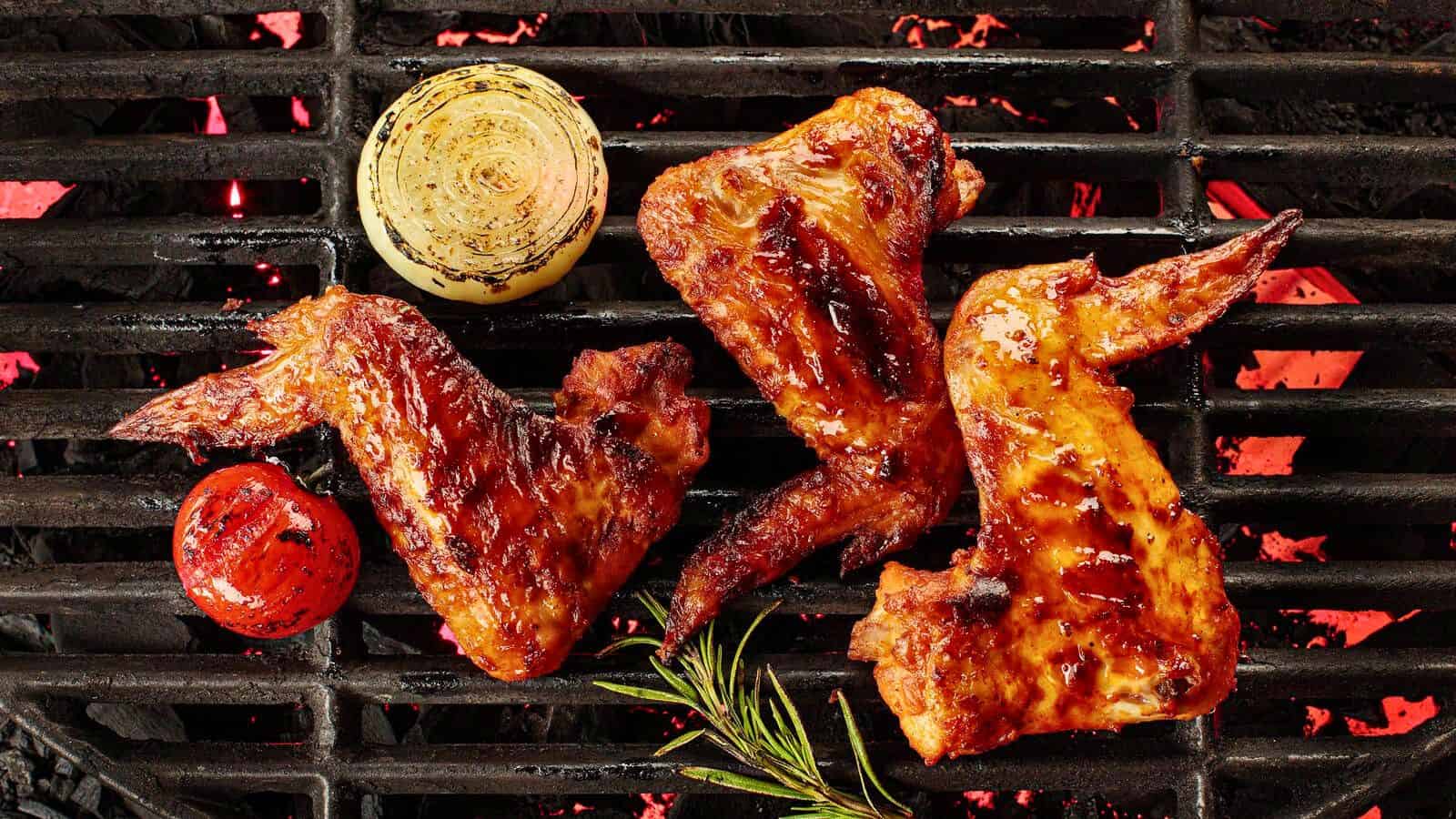 Three grilled chicken wings are cooking on a barbecue grill with a charred onion slice, a charred tomato, and a sprig of rosemary. The grill's bars have red-hot coal beneath, indicating it is in use. The chicken wings appear to be glazed.