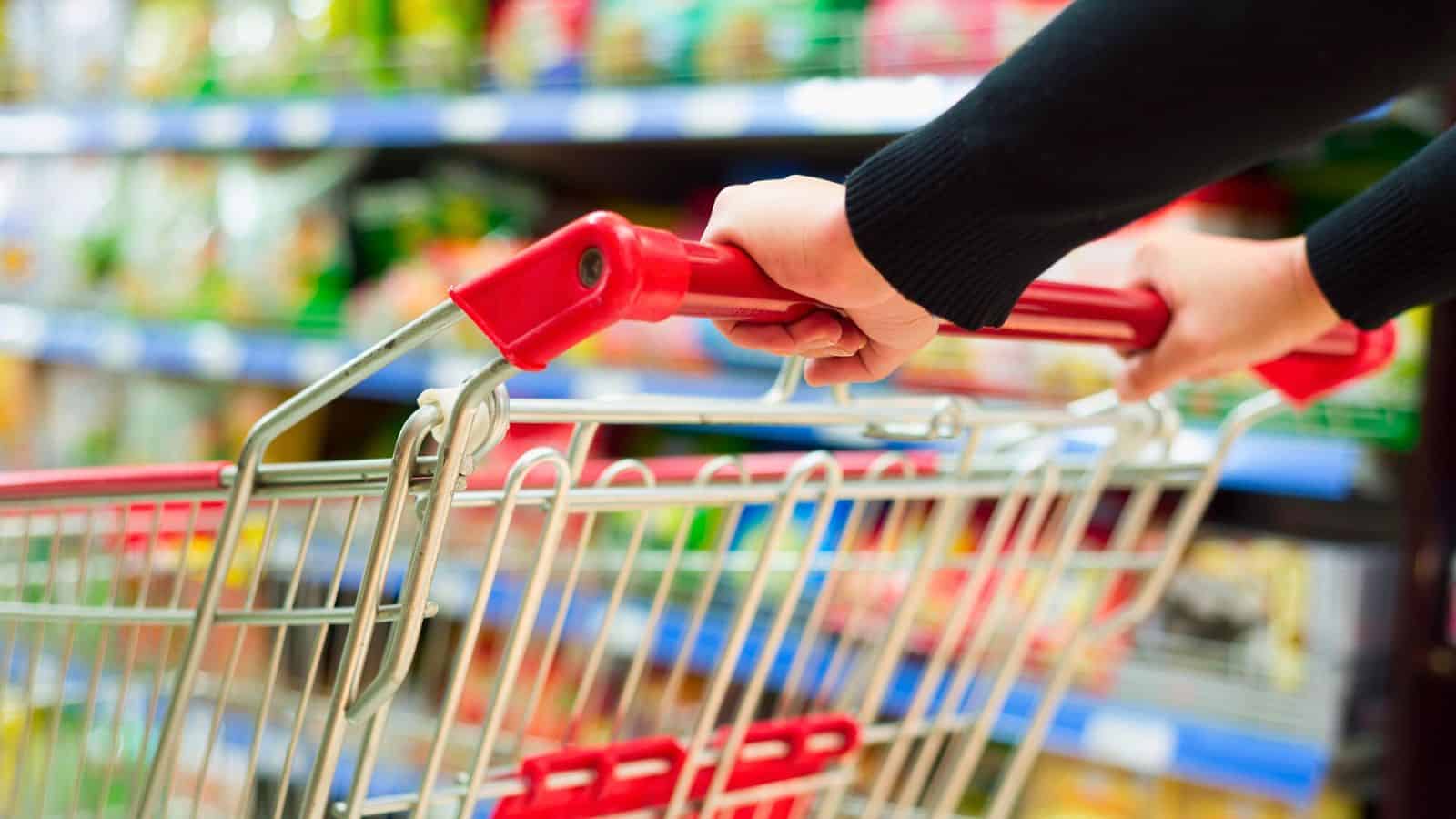 Close-up of hands pushing a shopping cart with a red handle in a grocery store aisle. Shelves stocked with various products are blurred in the background. The scene depicts typical grocery shopping activity.