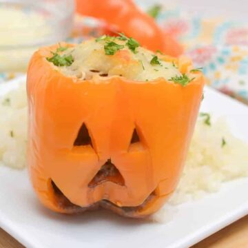 A stuffed orange bell pepper carved to resemble a jack-o'-lantern sits on a white plate in this Easy Halloween Recipe. The filling includes various ingredients, topped with parsley. The background shows a colorful napkin and a small bowl of grated cheese, perfect for festive Halloween dining.