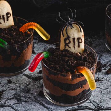 Three clear glasses filled with layers of brown mousse, topped with crumbled cookies, and decorated with gummy worms and cookie tombstones labeled "RIP."