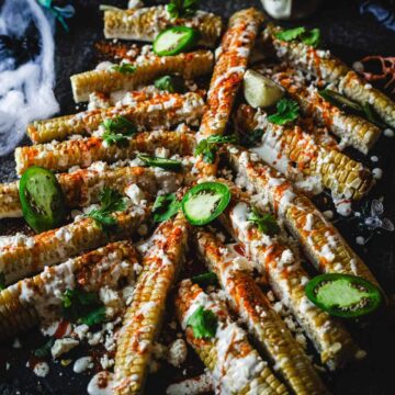 A Halloween-themed platter of corn ribs is shown. The corn ribs are seasoned with chili powder, topped with cheese, lime slices, jalapenos, and cilantro. The background features fake spider webs and a toy spider, adding to the spooky vibe—an easy Halloween recipe sure to thrill your guests!