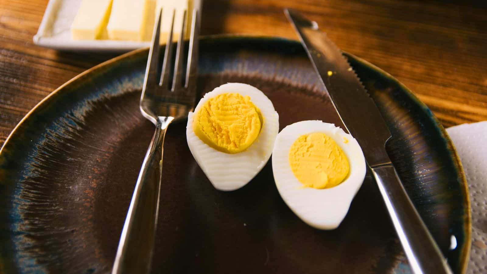 A halved hard-boiled egg is on a dark ceramic plate with a fork and knife on either side. In the background, a butter dish with butter slices is partially visible. The table is wooden, and a napkin is at the edge of the image.