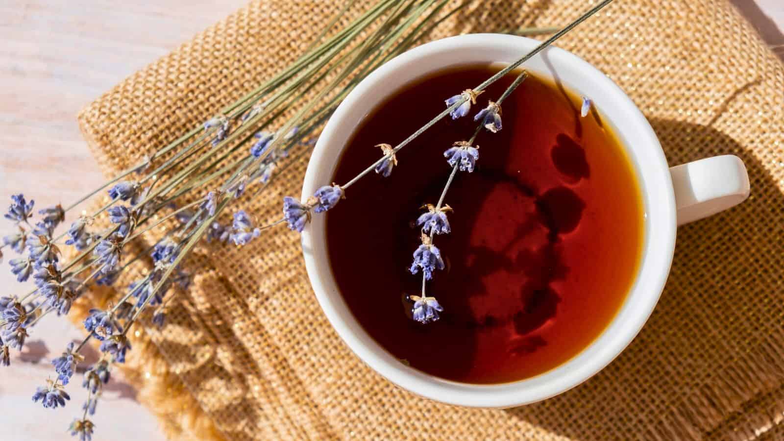 A white cup filled with dark tea is placed on a woven fabric surface. Stalks of small, delicate purple flowers are laid across the rim of the cup and the fabric underneath. The background is a light wooden texture.
