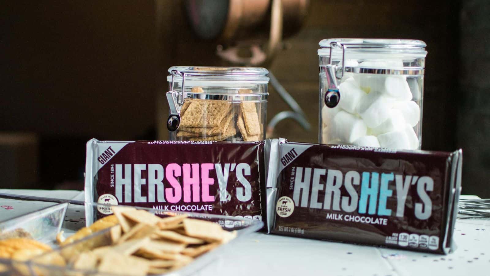 A table displays ingredients for making s'mores, including two large Hershey's milk chocolate bars, a container with graham crackers, another with large marshmallows, and some loose graham crackers on a plate.