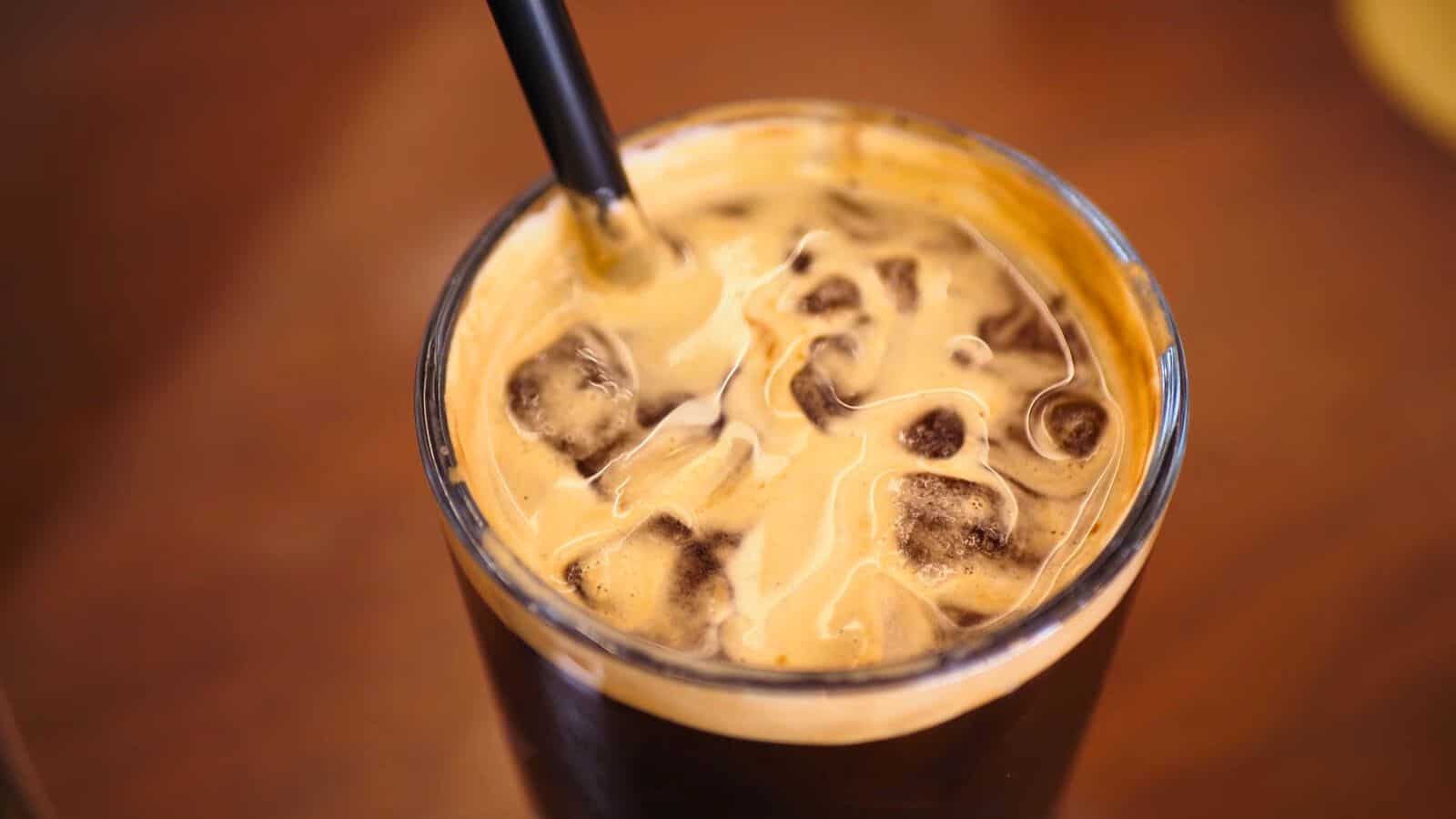 A close-up photo of an iced coffee in a glass. The coffee's surface is topped with a light froth and ice cubes, and a black straw is inserted into the drink. The background is blurred.