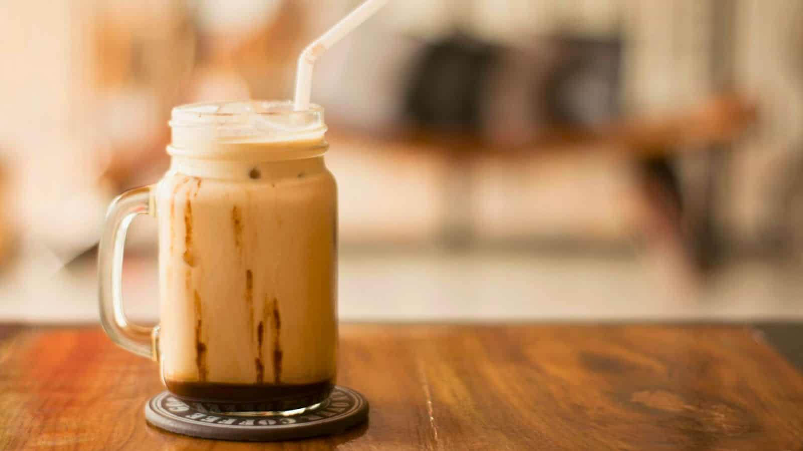 A glass mug filled with iced coffee, topped with foam, and a clear straw, sits on a coaster on a wooden table. The background is blurred and appears to be inside a casual dining area or cafe.