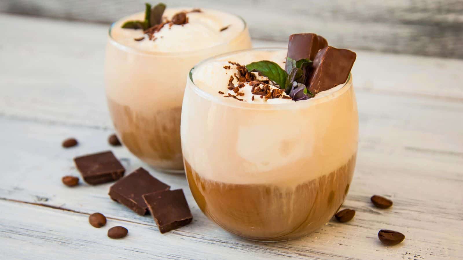 Two glass cups of a layered iced coffee beverage, topped with whipped cream, chocolate shavings, and mint leaves. Pieces of dark chocolate and coffee beans are scattered on the white wooden surface beside the drinks.