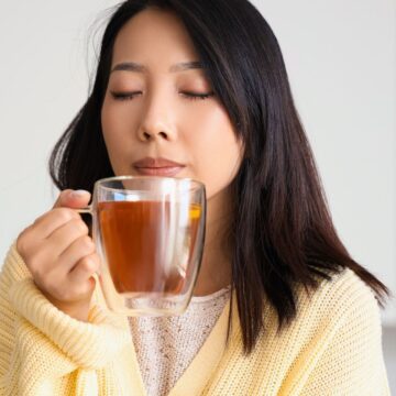 A person with long dark hair, wearing a light yellow sweater, is holding a glass cup filled with tea close to their face with eyes closed, appearing to enjoy the aroma. The background is softly blurred.