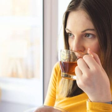 A person with long brown hair and a yellow sweater is drinking a dark-colored beverage from a glass mug. The individual is looking out of a window and holding the mug with both hands. The background shows a blurry indoor setting with natural light coming in.