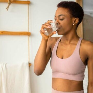 A woman dressed in a beige sports bra and athletic leggings stands indoors, drinking a glass of water. Behind her, there is a ladder shelf with towels and a large framed beach photograph on the wall.