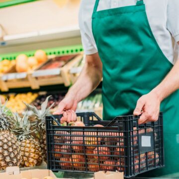 A person wearing a green apron and white shirt is carrying a black plastic basket filled with produce. In the background, another person selects fruits and vegetables from a display in a grocery store.