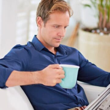 A man in a blue shirt sits on a white couch, holding a light blue mug in his right hand and reading a newspaper with his left hand. A potted plant is in the background.